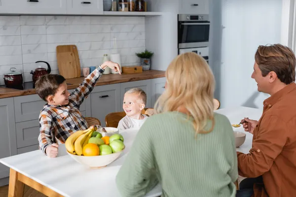 Niño señalando al hermano durante el desayuno con los padres cerca de un tazón de frutas frescas — Stock Photo