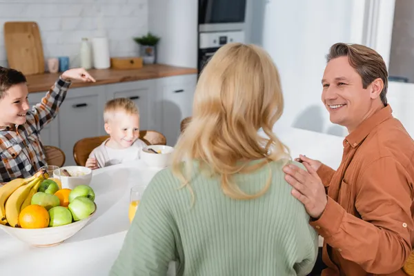 Vista trasera de la mujer rubia y el marido sonriente cerca de frutas frescas y niños desayunando - foto de stock