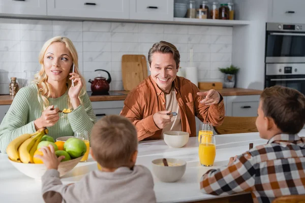 Вид сзади мальчиков рядом с матерью, говорящей по смартфону, и отцом, указывающим на апельсиновый сок во время завтрака — стоковое фото