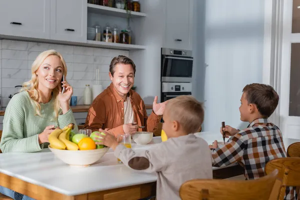 Fröhlicher Mann spricht während des Frühstücks mit Söhnen, während seine Frau mit dem Handy telefoniert — Stockfoto