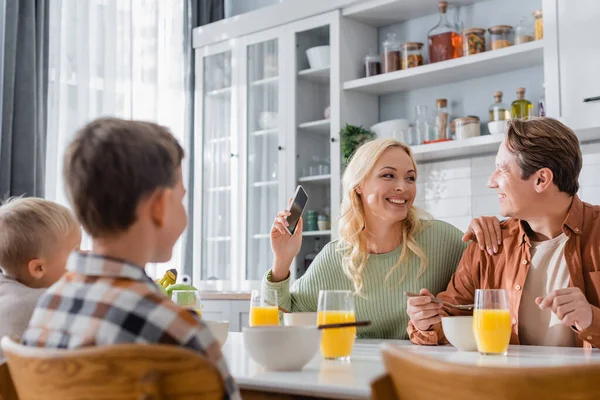 Радостная женщина с мобильным телефоном смотрит на мужа во время завтрака с размытыми детьми — стоковое фото