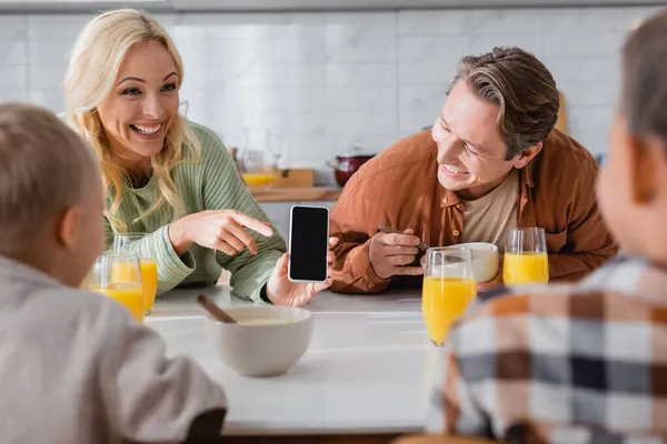 Весела жінка вказує на смартфон з порожнім екраном під час сніданку з чоловіком і розмитими синами — стокове фото