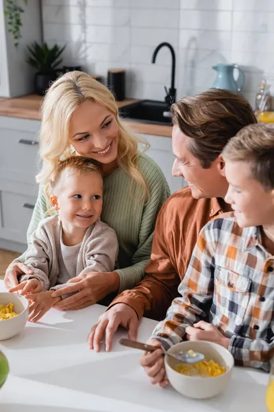 Счастливая семья смотрит друг на друга возле миски с кукурузными хлопьями на кухонном столе — стоковое фото