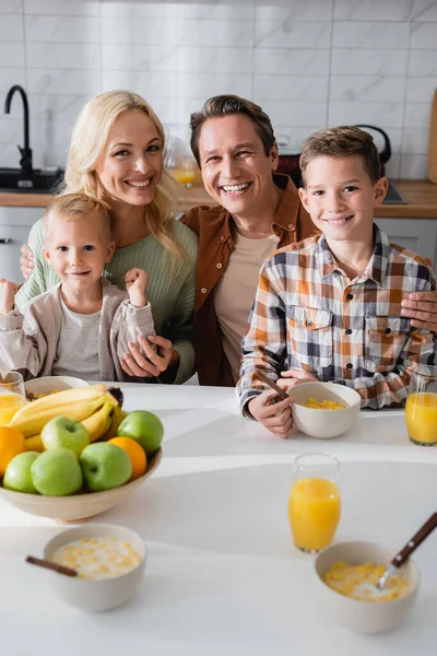 Casal alegre com filhos olhando para a câmera perto de frutas frescas e suco de laranja na cozinha — Fotografia de Stock