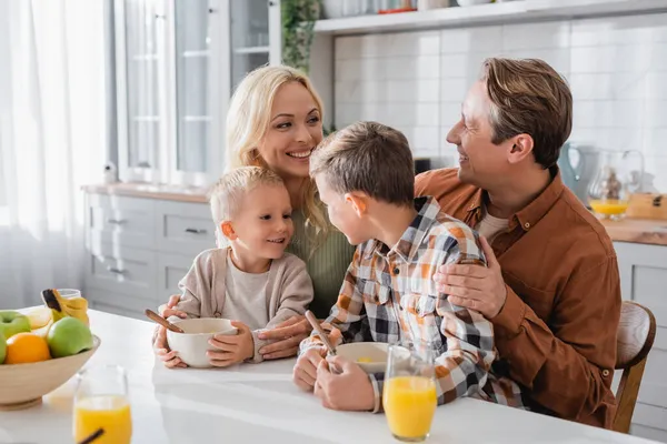 Feliz pareja mirándose y abrazando a sus hijos durante el desayuno - foto de stock
