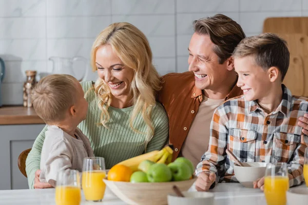 Радостная семья сидит за кухонным столом рядом со свежими овощами и апельсиновым соком на размытом переднем плане — стоковое фото