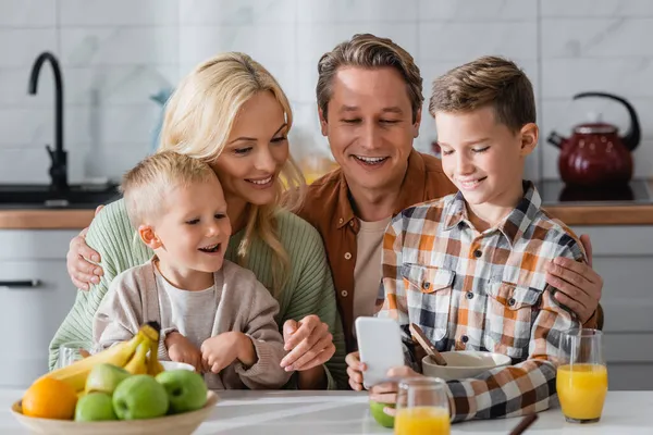 Niño sonriente mostrando teléfono inteligente a la familia feliz durante el desayuno - foto de stock