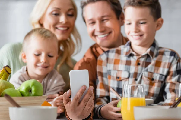 Homme prenant selfie sur smartphone près de famille heureuse floue pendant le petit déjeuner — Photo de stock