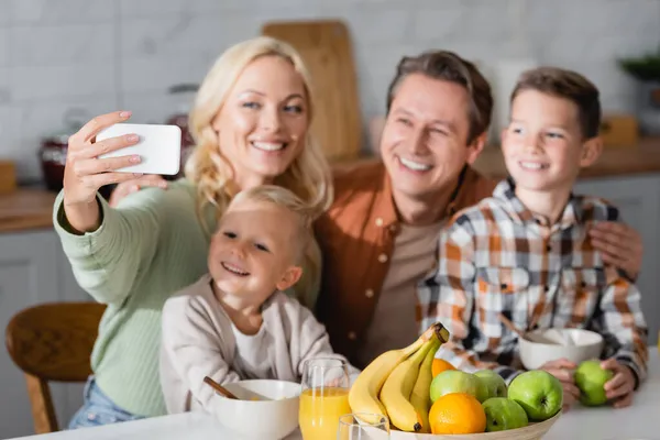 Mujer alegre tomando selfie en el teléfono celular con la familia cerca de frutas frescas durante el desayuno - foto de stock