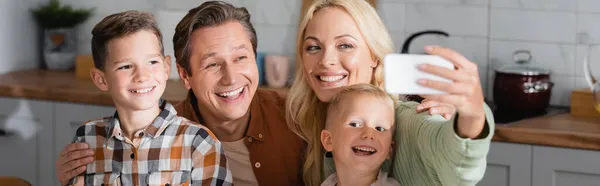 Mujer alegre tomando selfie en smartphone con el marido y los hijos en la cocina, pancarta - foto de stock