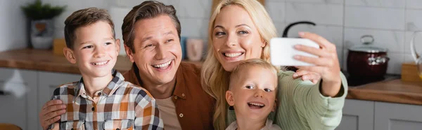 Femme gaie prenant selfie sur smartphone avec mari et fils dans la cuisine, bannière — Photo de stock