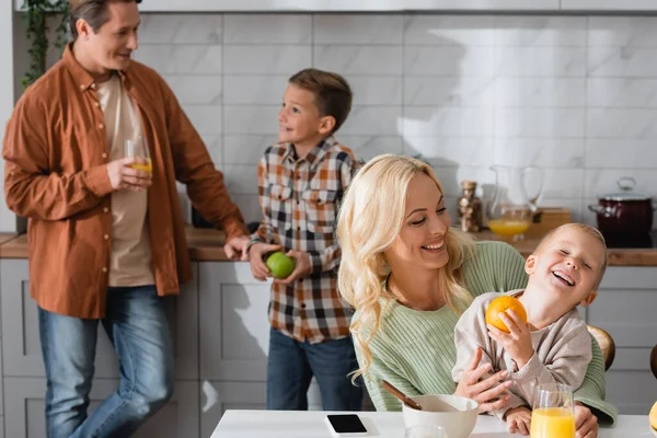 Mujer alegre divirtiéndose con el niño en la cocina cerca del padre hablando con el hijo sobre fondo borroso - foto de stock