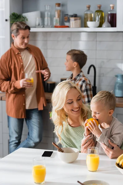 Довольный ребенок ест апельсин рядом с мамой и размытый папа с братом на кухне — стоковое фото