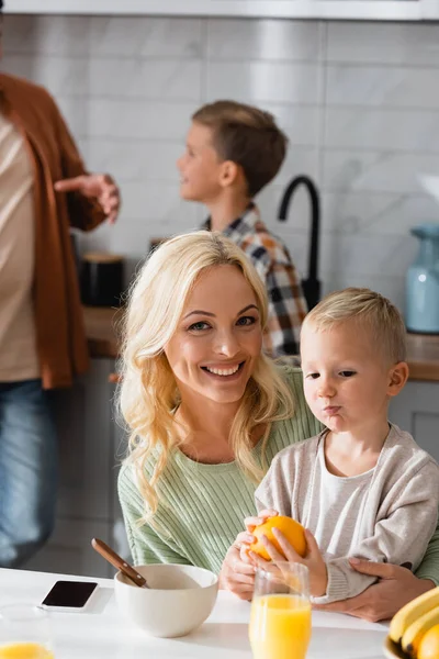 Happy woman looking at camera while holding kid with orange near father and son on blurred background — Stock Photo