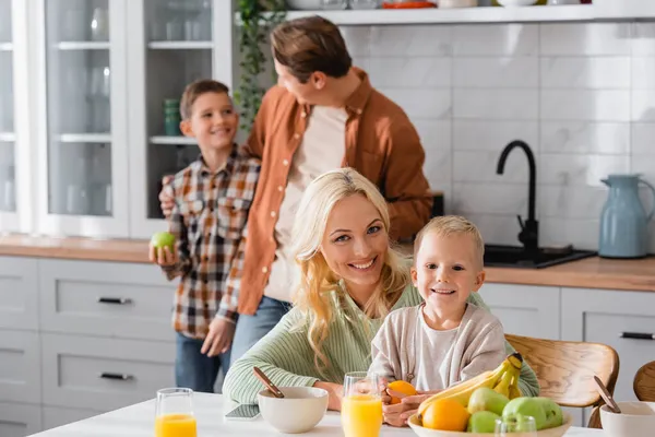 Mujer feliz mirando a la cámara mientras desayuna con el niño cerca borrosa marido e hijo - foto de stock