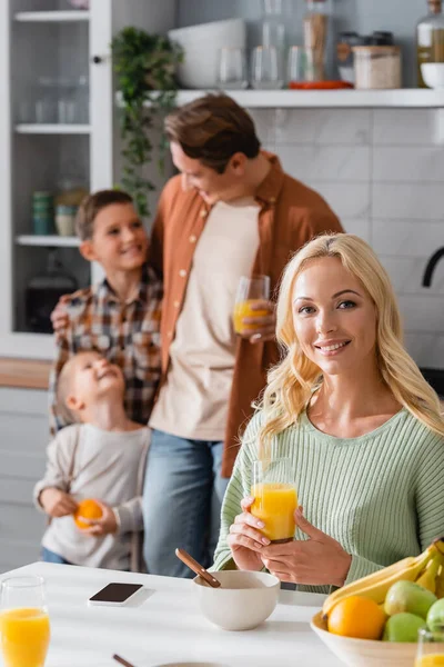 Femme gaie regardant caméra près flou mari et fils dans la cuisine — Photo de stock