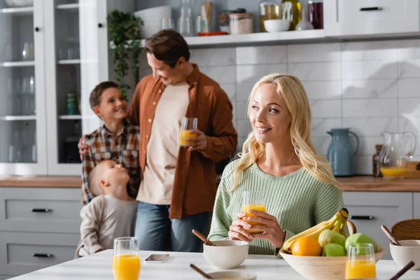 Donna gioiosa seduta vicino al tavolo con colazione vicino alla famiglia offuscata in cucina — Foto stock