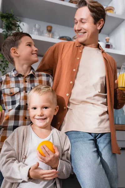 Happy boy holding orange near blurred dad and brother in kitchen — Stock Photo