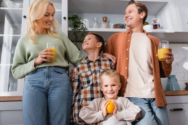 Feliz pareja sosteniendo vasos con jugo de naranja fresco cerca de los niños en la cocina - foto de stock