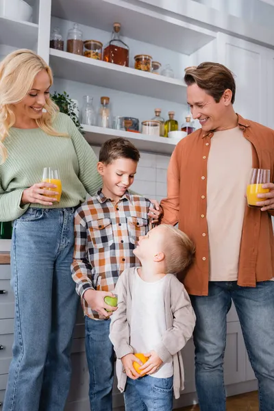 Улыбающаяся пара с апельсиновым соком, улыбающаяся рядом с сыновьями, держащими фрукты на кухне — стоковое фото