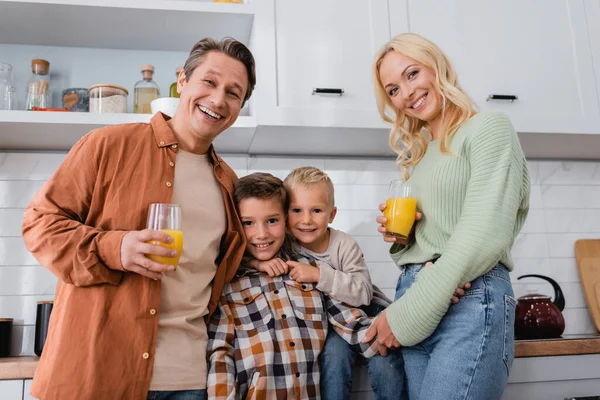 Irmãos alegres olhando para a câmera perto dos pais com suco de laranja na cozinha — Fotografia de Stock