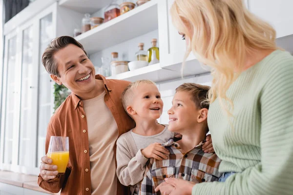 Homem alegre com suco de laranja olhando para filhos alegres e esposa na cozinha — Fotografia de Stock