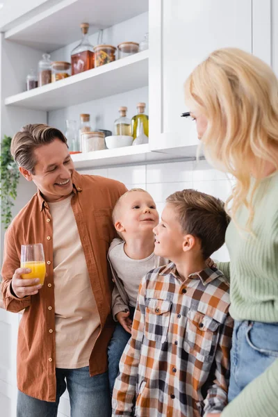 Hombre complacido sosteniendo jugo de naranja cerca de hijos felices y esposa borrosa en la cocina - foto de stock