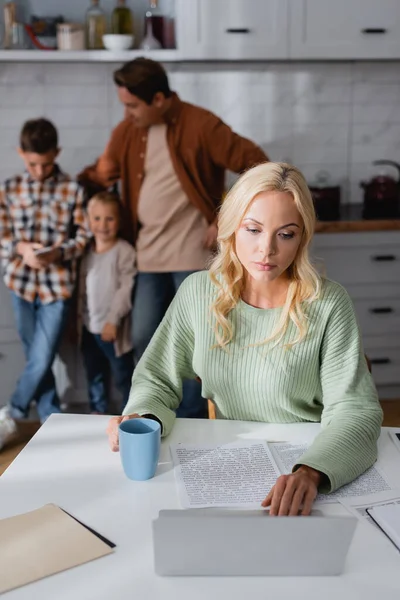 Busy mother working in kitchen near cup of tea and blurred husband with sons — Stock Photo