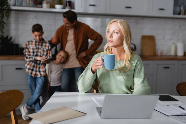 Thoughtful woman with cup of tea sitting near laptop, documents, and blurred husband with kids in kitchen — Stock Photo