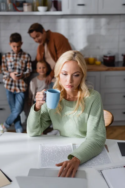 Woman with cup of tea working near documents and laptop near blurred family in kitchen — Stock Photo