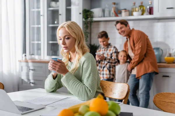 Busy woman with cup of tea working at laptop near blurred fruits and cheerful family in kitchen — Stock Photo