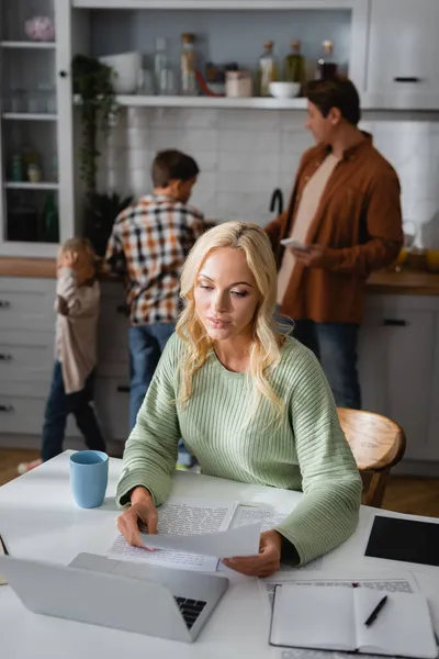 Mujer rubia que trabaja con documentos cerca de la computadora portátil en la cocina cerca del marido y los hijos en un fondo borroso - foto de stock