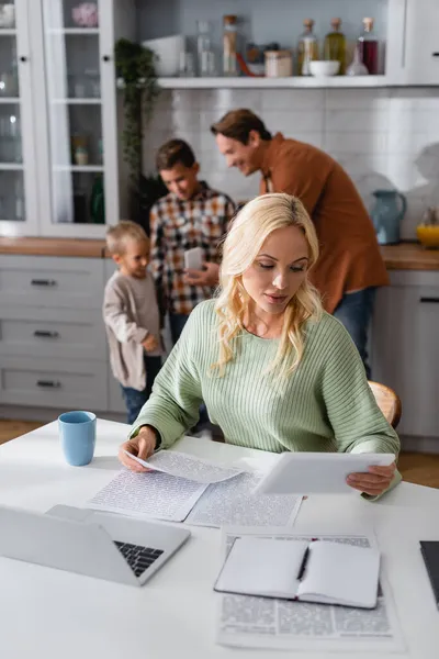 Mujer que trabaja con papeles y tabletas digitales limpios maridos borrosos e hijos en casa - foto de stock