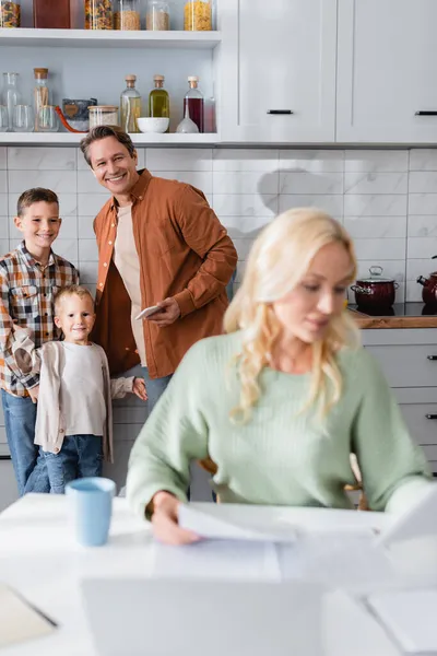 Mann mit Sohn lächelt neben beschäftigter Mutter, die in Küche in der Nähe von Dokumenten und verschwommenem Laptop arbeitet — Stockfoto