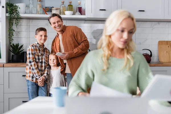 Hombre feliz con los niños cerca de la esposa que trabaja en la cocina en primer plano borrosa - foto de stock