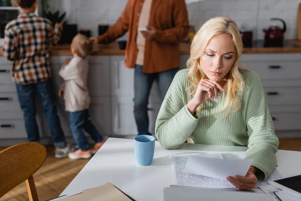 Femme sérieuse travaillant avec des documents près d'ordinateur portable flou et la famille dans la cuisine — Photo de stock