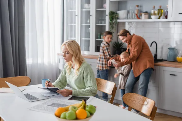 Busy woman writing in notebook while working near laptop and blurred family in kitchen — Stock Photo
