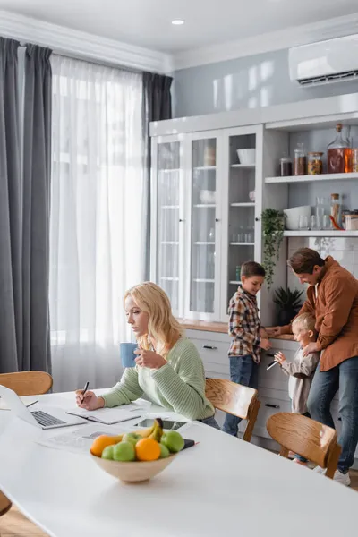 Woman with cup of tea writing in notebook near laptop and husband with sons in kitchen — Stock Photo