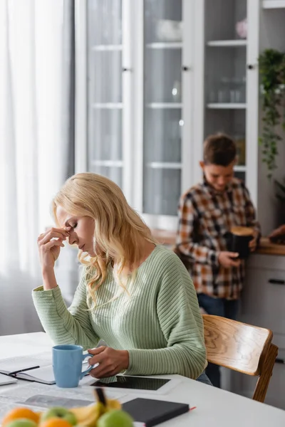 Tired woman with closed eyes sitting with cup of tea and digital tablet with blank screen  near blurred son in kitchen — Stock Photo