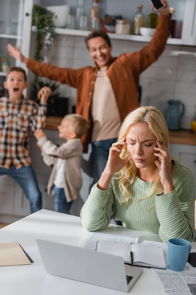 Mujer agotada sentada con los ojos cerrados cerca de la computadora portátil mientras la familia borrosa divertirse en la cocina - foto de stock