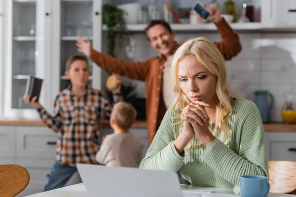 Famille floue avoir du plaisir dans la cuisine près femme réfléchie travaillant près d'un ordinateur portable — Photo de stock