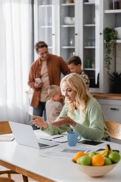 Woman pointing with hands during video call on laptop near blurred husband and sons in kitchen — Stock Photo