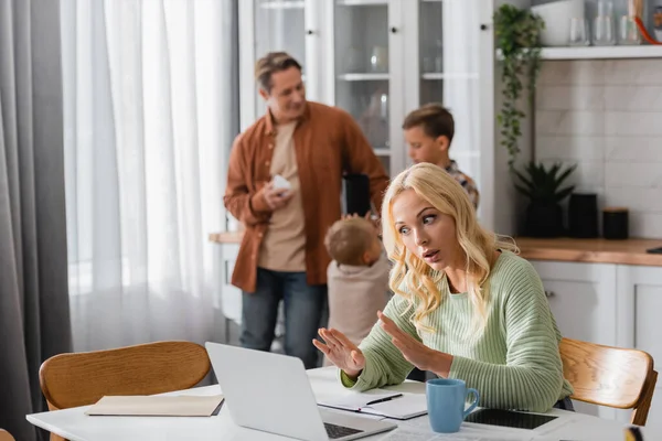 Frau zeigt Stopp-Geste bei Videoanruf auf Laptop in Küche nahe Familie auf verschwommenem Hintergrund — Stockfoto