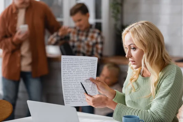 Frau zeigt bei Videoanruf auf Laptop neben verschwommener Familie in Küche auf Dokument — Stockfoto