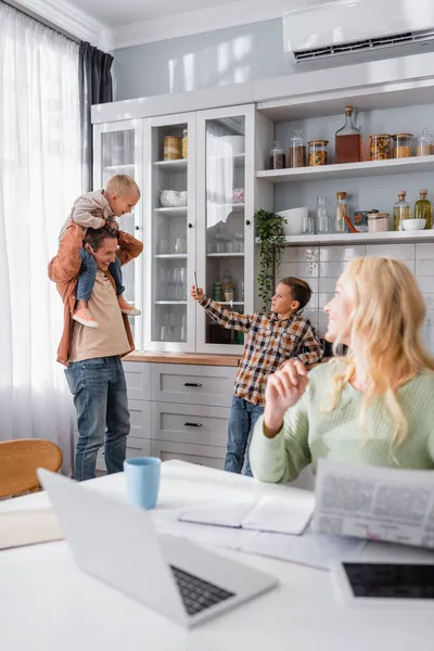 Мужчина играет с детьми на кухне рядом с женой работает рядом с ноутбуком на размытом переднем плане — стоковое фото
