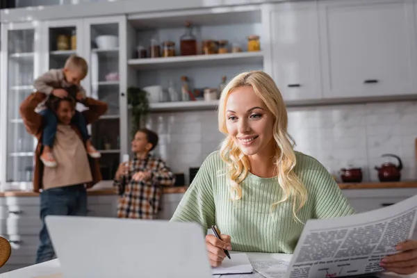 Mujer sonriente con periódico y pluma trabajando cerca de la computadora portátil y la familia borrosa en la cocina - foto de stock