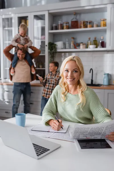 Mujer sonriente con la escritura del periódico en el cuaderno cerca de gadgets y la familia borrosa divertirse en la cocina — Stock Photo