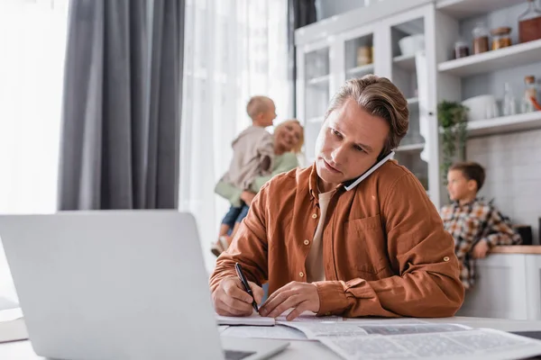 Homme parlant sur smartphone tout en travaillant près de la famille floue avoir du plaisir dans la cuisine — Photo de stock