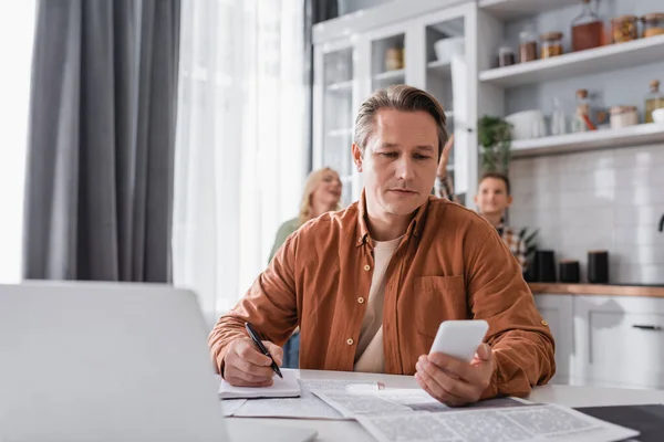 Man writing in notebook while looking at smartphone near laptop and blurred family in kitchen — Stock Photo
