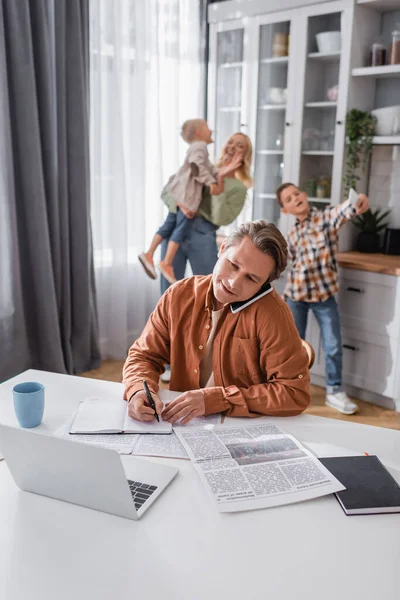 Mann spricht auf Smartphone und schreibt in Notizbuch, während er in der Küche in der Nähe einer verschwommenen Familie arbeitet — Stockfoto